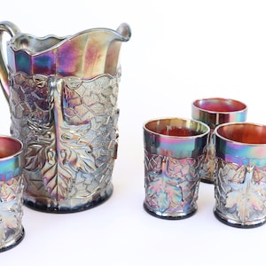 May include: A vintage iridescent glass pitcher and matching tumblers. The pitcher has a handle and is decorated with a leaf and vine pattern. The tumblers have a similar design. The glass has a metallic sheen with colors of purple, blue, and gold.
