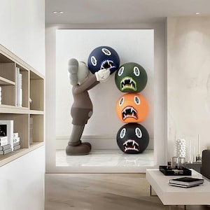 May include: A brown and black figurine with a large head and arms holding a stack of colourful balls with faces. The balls are stacked on top of each other and have different expressions. The background is a white wall.