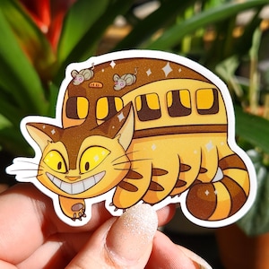 May include: A whimsical sticker featuring a cat-shaped bus with a smiling face and yellow eyes. The bus is yellow and brown with mouse passengers on top. The sticker has a white border and sparkling accents.