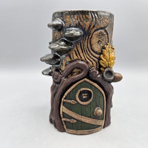 May include: A whimsical ceramic tree stump-shaped container with a fairy door, mushrooms, and foliage. The container is painted in shades of brown, green, and gold, creating a fantasy-themed decorative piece. The container is approximately 5 inches tall.