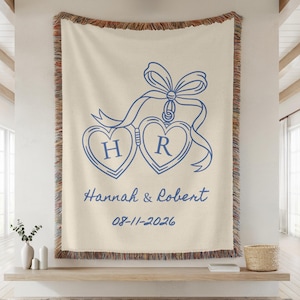 May include: Cream-colored woven throw blanket with a blue heart-shaped design, ribbon, and the initials "H" and "R". The blanket also features the names "Hannah & Robert" and the date "08-11-2026".