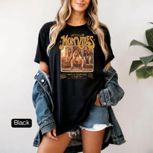 May include: Black t-shirt with a graphic that reads "Mom Vibes" in gold lettering. The graphic features a sepia-toned image of women. The text "raised on cassette tapes & tough love" is also on the shirt.