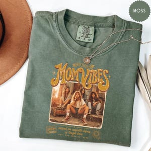 May include: A moss green Comfort Colors t-shirt with the text "Mom Vibes" in a retro design. The graphic features four women. A silver necklace with a pendant rests on the shirt. The shirt also has the text "raised on cassette tapes & tough love".