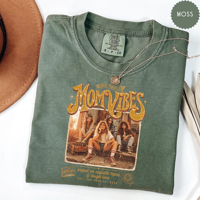 May include: A moss green Comfort Colors t-shirt with the text "Mom Vibes" in a retro design. The graphic features four women. A silver necklace with a pendant rests on the shirt. The shirt also has the text "raised on cassette tapes & tough love".