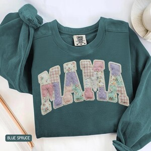 May include: A teal sweatshirt with the word "MAMA" appliqued across the chest. The letters are made of patchwork fabric in floral and plaid patterns, outlined with small white beads. The bottom left corner of the sweatshirt has the text "BLUE SPRUCE".