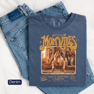 May include: A denim blue t-shirt with the words "Mom Vibes" in a retro design, featuring a group of women. The shirt also has the text "raised on cassette tapes & tough love" and "EST. THE GOOD OL' DAYS". The shirt is displayed on a pair of blue jeans.