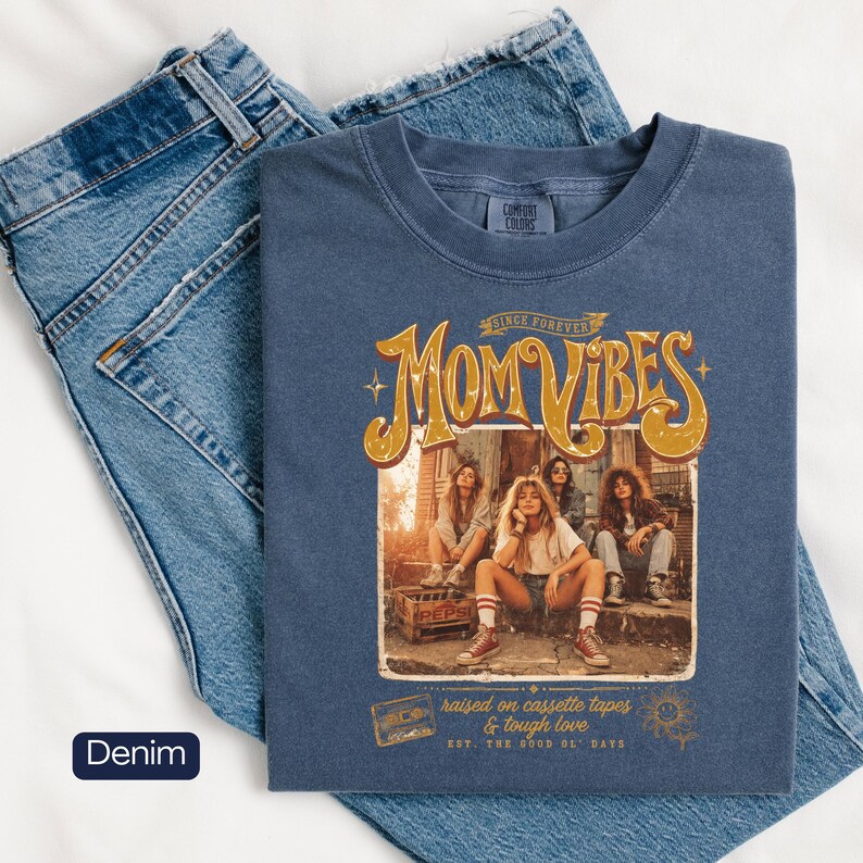 May include: A denim blue t-shirt with the words "Mom Vibes" in a retro design, featuring a group of women. The shirt also has the text "raised on cassette tapes & tough love" and "EST. THE GOOD OL' DAYS". The shirt is displayed on a pair of blue jeans.