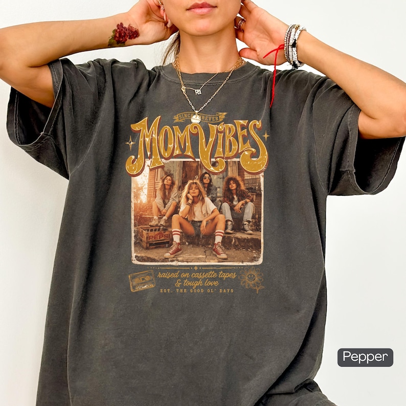 May include: Dark gray t-shirt with the words "MOM VIBES" in a retro font. The graphic features a vintage photo of four women. The text "raised on cassette tapes & tough love" is also printed on the shirt.