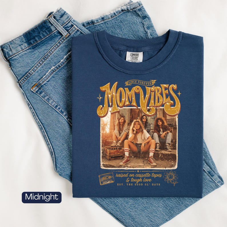 May include: Midnight blue sweatshirt with the text "MOM VIBES" in gold lettering, featuring a vintage-style graphic of four women. The sweatshirt is paired with light blue denim jeans. The text "raised on cassette tapes & tough love" is also visible.