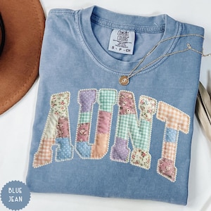 May include: A blue jean Comfort Colors t-shirt with the word "AUNT" appliqued in patchwork fabric. The patchwork letters are outlined with small white beads. A gold necklace with a pendant is on the shirt. The shirt has a small tag that says "S P CH".