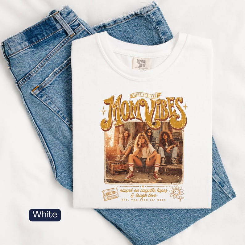 May include: White t-shirt with the text "MOM VIBES" in a retro design, featuring a group of women. The shirt also includes the phrases "raised on cassette tapes & tough love" and "the good ol' days". The shirt is displayed on top of blue jeans.
