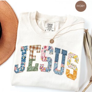 May include: Ivory-colored t-shirt with the word "JESUS" appliquéd in various patterned fabrics. A gold necklace with a pendant rests on the shirt. The shirt is from Comfort Colors and is a size small.