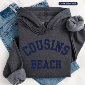 May include: Dark heather gray hooded sweatshirt with the words "COUSINS BEACH" in navy blue block letters. The sweatshirt is folded on top of a pair of blue jeans. The hoodie has a drawstring and a soft, fleece lining.