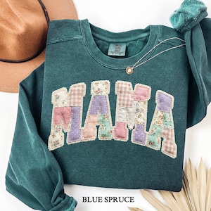 May include: A dark teal long-sleeve crewneck sweatshirt featuring the word MAMA in large, block-style letters. Each letter is filled with a patchwork of floral, gingham, and polka dot fabric patterns in pastel pink, purple, and green tones.