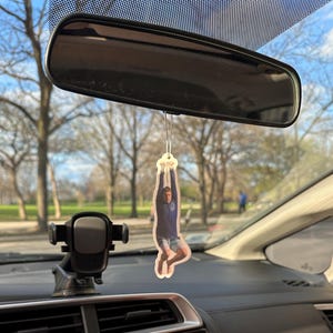 May include: A car air freshener featuring a photo of a person hanging upside down from a rope. The person is wearing a blue shirt and shorts.