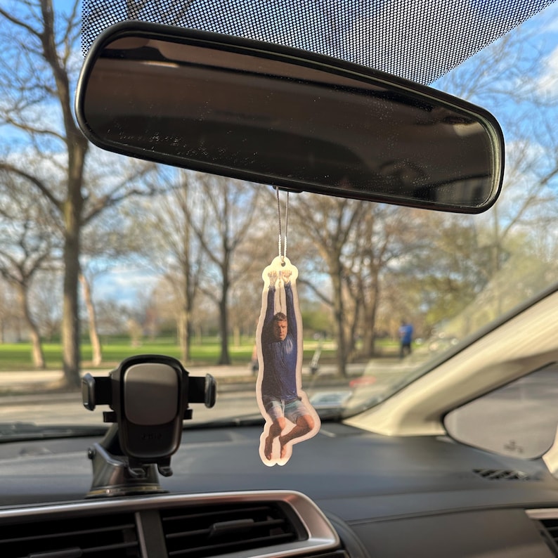 May include: A car air freshener featuring a photo of a person hanging upside down from a rope. The person is wearing a blue shirt and shorts.