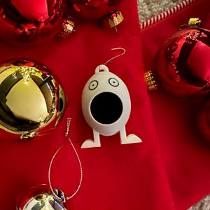 May include: White ceramic Christmas ornament shaped like a surprised face with a large open mouth. The ornament is hanging from a red velvet bag with other ornaments.