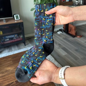 Puede incluir: Un calcetín negro con un patrón geométrico en azul, amarillo y morado. El calcetín tiene talón y puntera negros. El diseño presenta líneas y formas de media luna.