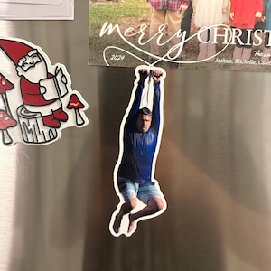 Puede incluir: Un imán de nevera con un hombre con una camisa azul de manga larga y pantalones cortos estampados, colgado de una cuerda. El imán está en la puerta plateada de una nevera, cerca de un imán de gnomo y seta y una tarjeta de Navidad.