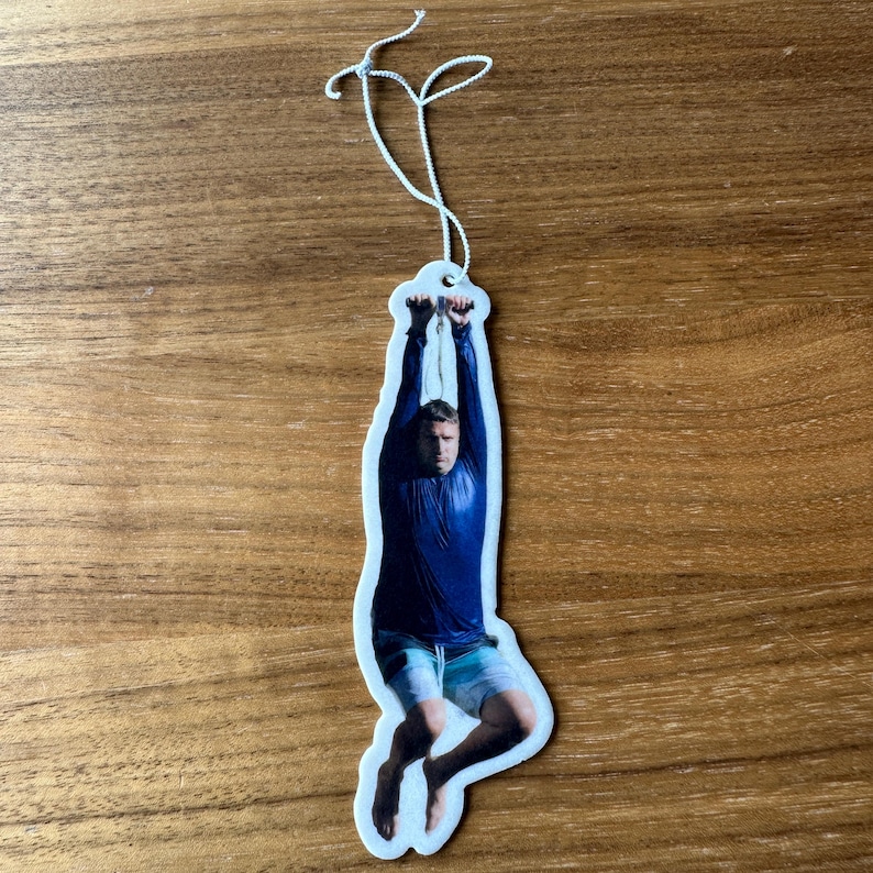 May include: A white car air freshener with a photo of a person hanging from a rope. The person is wearing a blue shirt and shorts.