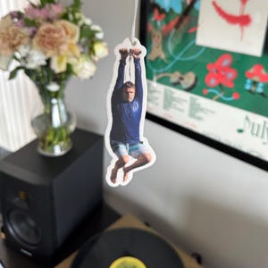 May include: A white car air freshener with a photo of a person hanging from a rope. The person is wearing a blue shirt and blue shorts.
