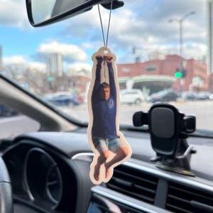 May include: A white car air freshener with a photo of a person hanging upside down. The person is wearing a blue shirt and shorts.