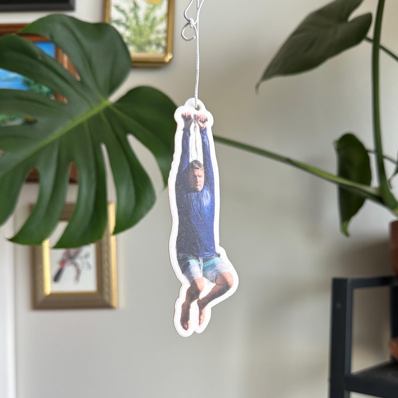 May include: A white car air freshener with a photo of a person hanging from a rope. The person is wearing a blue shirt and shorts.