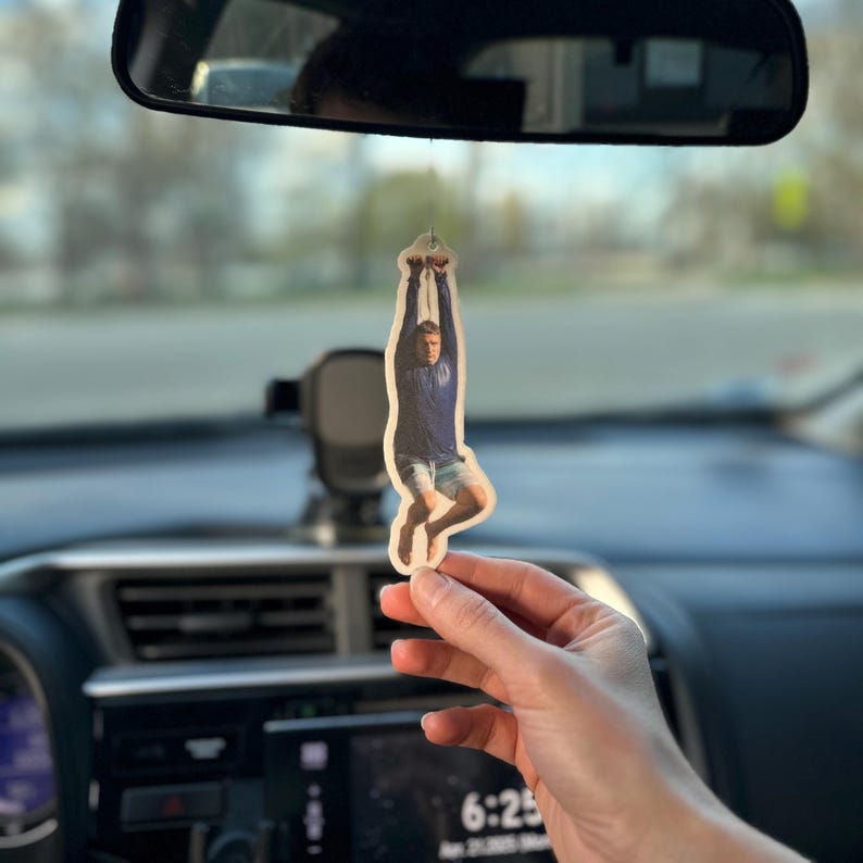 May include: A car air freshener featuring a photo of a person hanging upside down from a rope. The person is wearing a blue shirt and shorts.