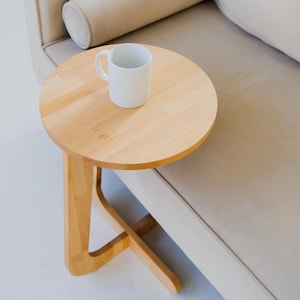 May include: A small, round, light-colored wooden side table with a white mug on top. The table has a unique, curved leg design and is positioned next to a beige sofa. The table's top is approximately 18 inches in diameter.
