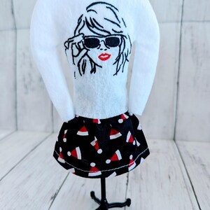 May include: A white long-sleeved shirt with an embroidered image of a woman wearing sunglasses. The shirt is paired with a black skirt with a red and white Santa hat pattern.