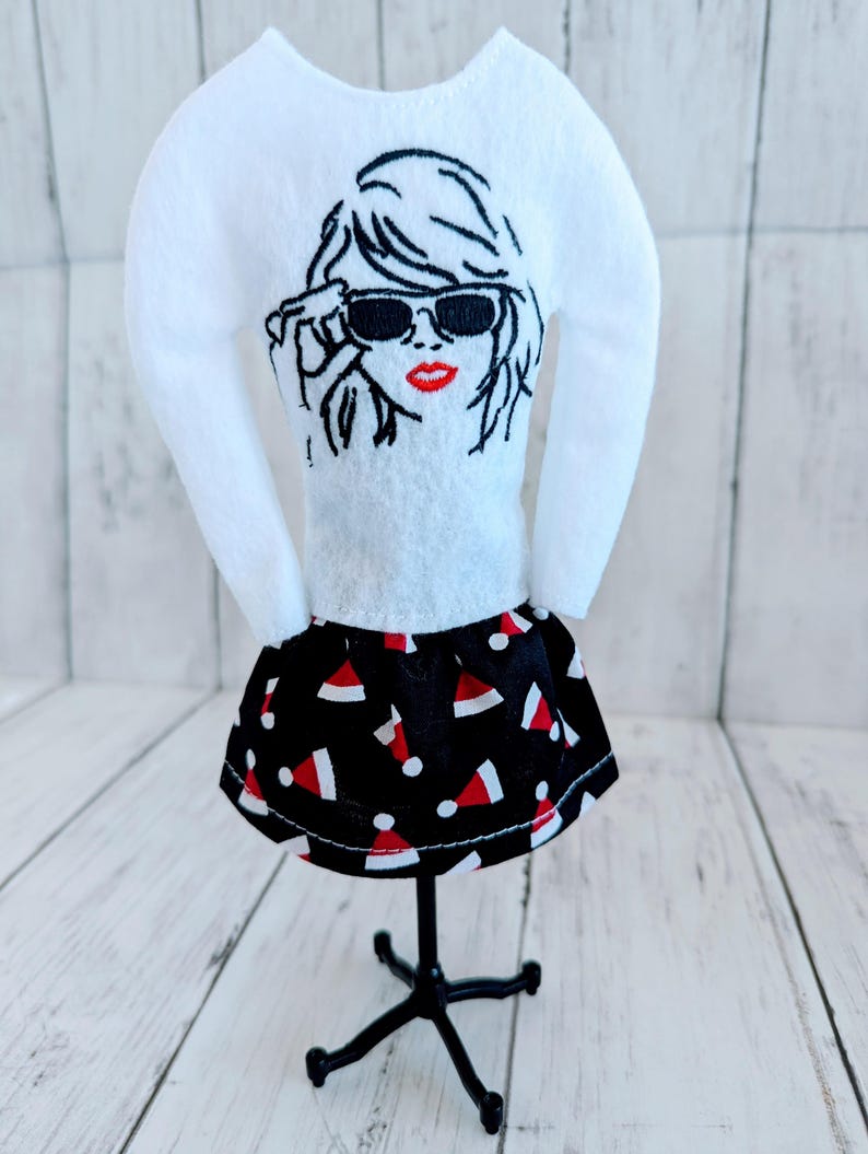 May include: A white long-sleeved shirt with an embroidered image of a woman wearing sunglasses. The shirt is paired with a black skirt with a red and white Santa hat pattern.