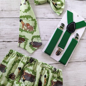 May include: A green and brown baby outfit with a jungle theme. The outfit includes a bow tie, suspenders, and a diaper cover. The diaper cover features a pattern of jungle animals, including monkeys, snakes, and elephants. The bow tie and suspenders are also green and brown.