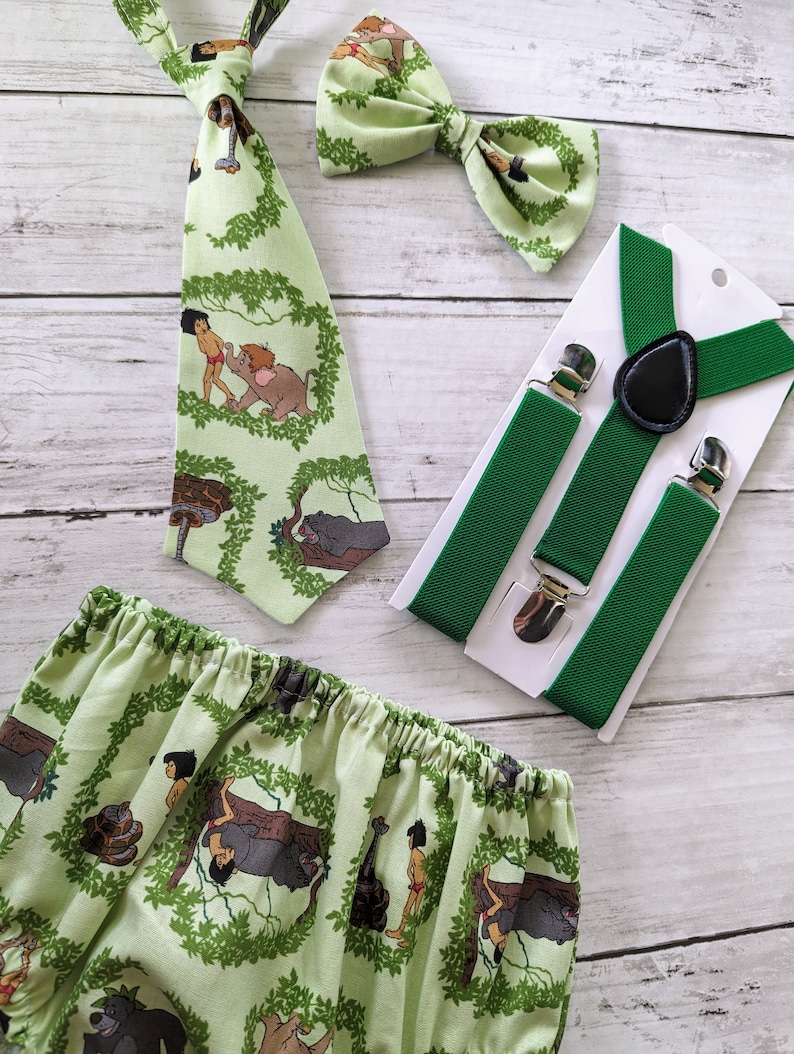 May include: A green and brown baby outfit with a jungle theme. The outfit includes a bow tie, suspenders, and a diaper cover. The diaper cover features a pattern of jungle animals, including monkeys, snakes, and elephants. The bow tie and suspenders are also green and brown.