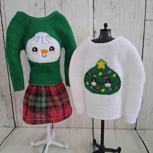 May include: A green long-sleeve felt shirt with a white dumpling graphic and a red and green plaid skirt on a white stand, next to a white long-sleeve felt shirt with a green Christmas tree graphic on a black stand for doll clothing.