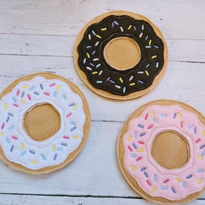 May include: Three felt donut coasters with different colored frosting and sprinkles. The donuts are brown, white, and pink.