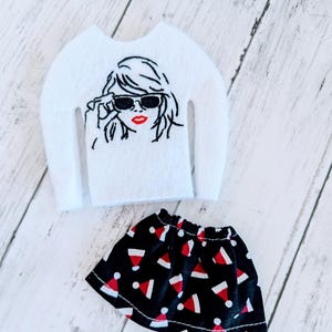 May include: A white felt sweater with a black and white embroidered image of a woman wearing sunglasses and red lipstick. A black skirt with a red and white Santa hat pattern.