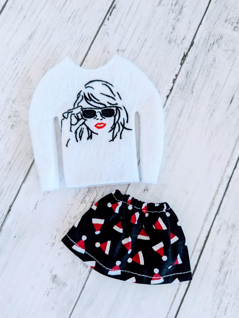 May include: A white felt sweater with a black and white embroidered image of a woman wearing sunglasses and red lipstick. A black skirt with a red and white Santa hat pattern.