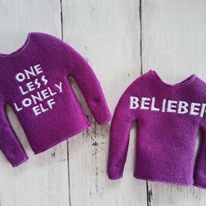 May include: Two purple felt sweaters with white text. One says "ONE LESS LONELY ELF" and the other says "BELIEBER". The sweaters have long sleeves and a rounded neckline. They are likely for an elf doll.