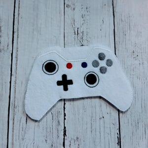 May include: White felt video game controller with black, red, and gray details. The controller features a directional pad, buttons, and analog sticks. The background is a light wood grain.