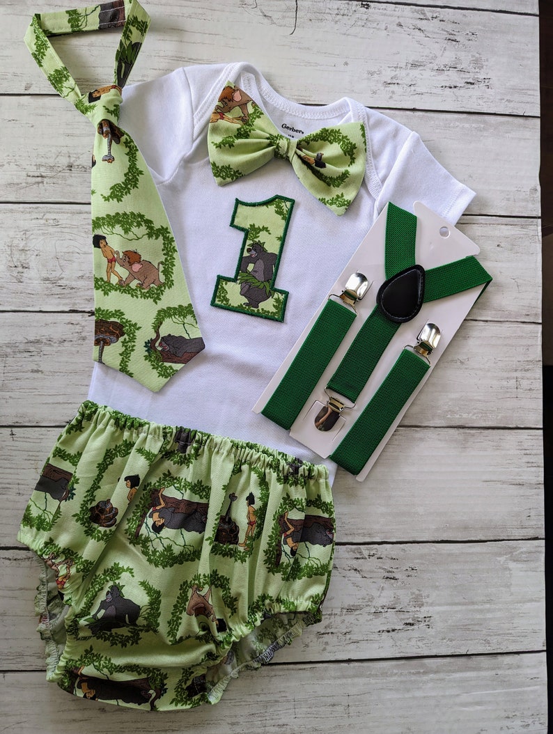 May include: A baby outfit for a first birthday party, featuring a white onesie with a green bow tie, a green and brown tie, a green and brown bow tie, a green and brown diaper cover, and a pair of green suspenders. The onesie has a green number one with a jungle scene on it. The diaper cover has a jungle scene with monkeys, snakes, and vines.