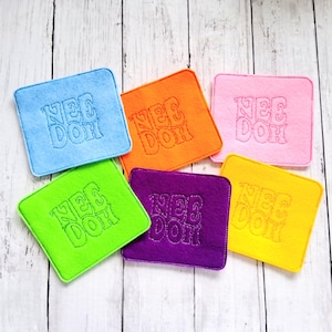 May include: Six square felt coasters in blue, orange, pink, lime green, purple, and yellow. Each coaster features the text "NEE DOH" embroidered in the center with a matching thread color, creating a textured look on the soft fabric surface.