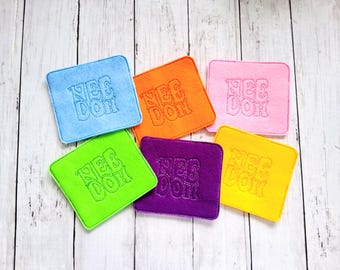 Könnte beinhalten: Sechs quadratische Filzuntersetzer in Blau, Orange, Pink, Limettengrün, Lila und Gelb. Jeder Untersetzer ist in der Mitte mit dem Text "NEE DOH" in passender Garnfarbe bestickt, was eine strukturierte Optik auf der weichen Oberfläche erzeugt.