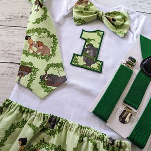 May include: A green and white baby outfit with a jungle theme. The outfit includes a white bodysuit with a green bow tie, a green tie with a jungle print, a green and white bow tie, a green and white skirt, and green suspenders. The bodysuit has a green felt number one with a jungle print.