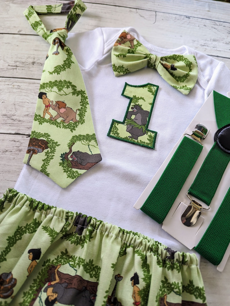 May include: A green and white baby outfit with a jungle theme. The outfit includes a white bodysuit with a green bow tie, a green tie with a jungle print, a green and white bow tie, a green and white skirt, and green suspenders. The bodysuit has a green felt number one with a jungle print.