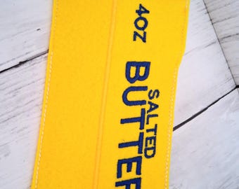 Könnte beinhalten: Ein gelber Filzausschnitt in Form eines Butterstücks mit weiß vernähten Kanten. Der Text 4oz SALTED BUTTER ist in dunkelblauem Garn auf die Vorderseite gestickt. Das Design weist eine vertikale Nahtlinie in der Mitte des gelben Filzes auf.
