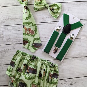 May include: A green and white baby outfit with a bow tie, suspenders, and a tie featuring characters from the Jungle Book. The outfit is made of cotton and is perfect for a baby boy.