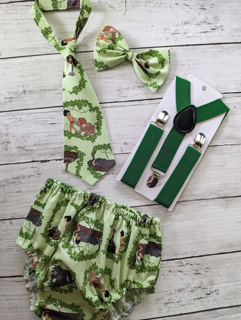 May include: A green and white baby outfit with a bow tie, suspenders, and a tie featuring characters from the Jungle Book. The outfit is made of cotton and is perfect for a baby boy.