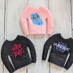 May include: Three miniature long-sleeved shirts. One is pink with a blue owl graphic, and two are black. One black shirt has red text that says "Saja Boys", and the other has a blue and white graphic with the text "Hunter".