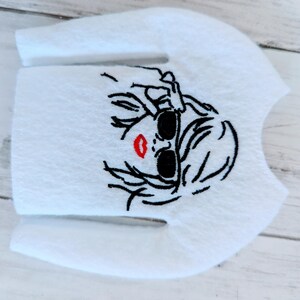 May include: White long-sleeved shirt with an embroidered design of a woman wearing sunglasses and red lipstick.