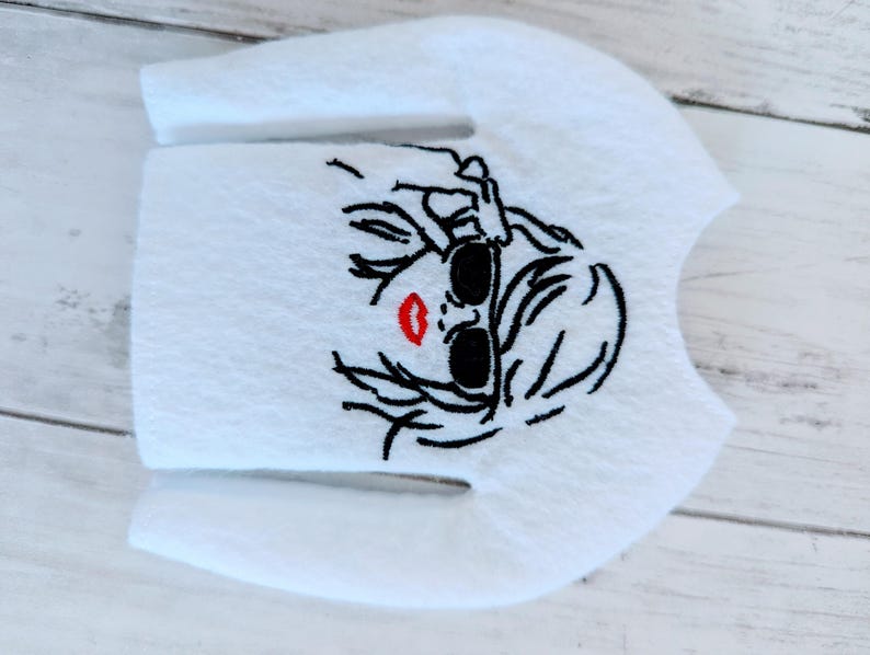 May include: White long-sleeved shirt with an embroidered design of a woman wearing sunglasses and red lipstick.